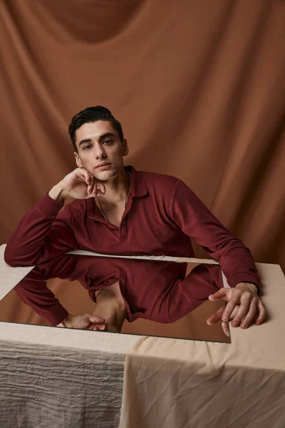 Όμορφος τύπος με πουκάμισο που ποζάρει σε τραπέζι σε εσωτερικό χώρο με φόντο ύφασμα και τετράγωνο καθρέφτη. — Φωτογραφία Αρχείου