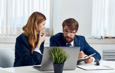 Bir kadın ve takım elbiseli bir adam dizüstü bilgisayara ve arka plandaki saksıdaki çiçeklere bakar.