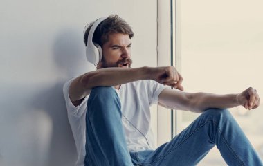 Bir adam pencerenin kenarına oturmuş kulaklık takıyor ve müzik dinliyor.
