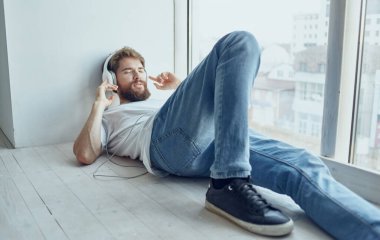 Neşeli sakallı adam, kulaklıkla pencere eşiğinde müzik teknolojisiyle yatıyor.