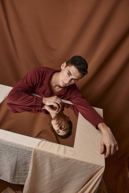 Kare aynalı bir adam kumaş arka planda ve kahverengi gömlekli bir masada oturur.