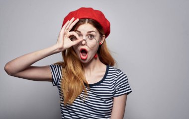 Tişörtlü güzel bir bayan, el hareketi yapıyor, gözlüklü, kırmızı şapkalı.