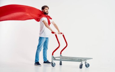 Kırmızı pelerinli sakallı bir adam elinde bir kargo arabası tutuyor.
