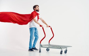 Kırmızı pelerinli sakallı bir adam elinde bir kargo arabası tutuyor.
