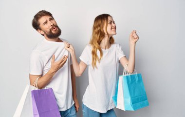 Beyaz tişörtlü genç bir çift ellerinde paketlerle neşe içinde alışveriş yapıyorlar.