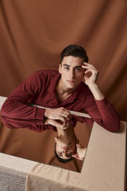 Masada aynası olan bir adam ormanın yakınında elini tutuyor ve kahverengi bir gömlek giyiyor.