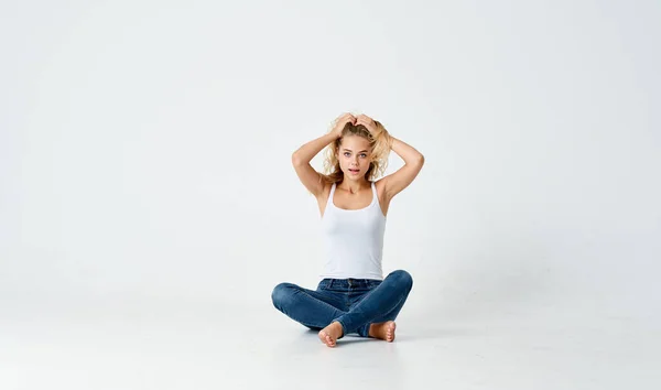 Donna in jeans e t-shirt bianca tocca i capelli sulla testa e si siede sul pavimento al chiuso — Foto Stock
