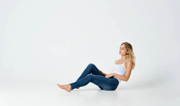 Bela loira em jeans e uma camiseta branca senta-se no chão dentro de casa — Fotografia de Stock