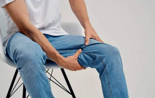 Hombre en jeans sentado en una silla y tocando su pierna con las manos recortadas vista — Foto de Stock