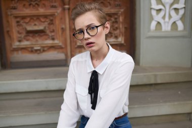 Kotlu ve gömlekli bir öğrenci binanın yanındaki basamaklarda oturuyor ve yüzünde gözlük var.