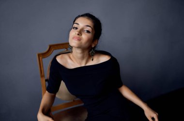 Siyah elbiseli zarif kadın gri odadaki tahta koltukta oturuyor.