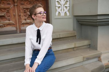 Kotlu ve gömlekli bir öğrenci binanın yanındaki basamaklarda oturuyor ve yüzünde gözlük var.