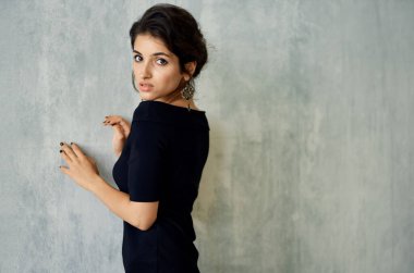 Gece elbisesi giymiş romantik bir esmer. Kapalı alanda gri bir duvara yaslanmış. Küpeler makyaj modeli.