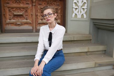 Kotlu ve gömlekli bir öğrenci binanın yanındaki basamaklarda oturuyor ve yüzünde gözlük var.