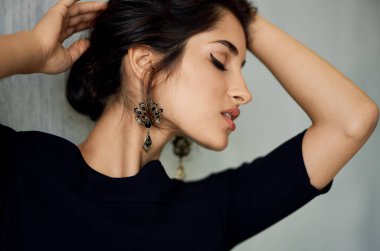 Gece elbisesi giymiş romantik bir esmer. Kapalı alanda gri bir duvara yaslanmış. Küpeler makyaj modeli.