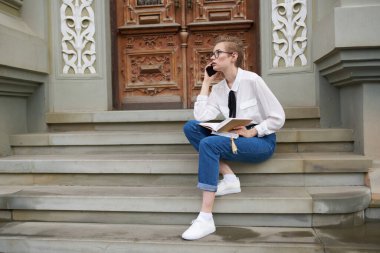 Bayan öğrenci elinde bir kitap ve cep telefonuyla binanın yanındaki basamaklarda oturuyor.