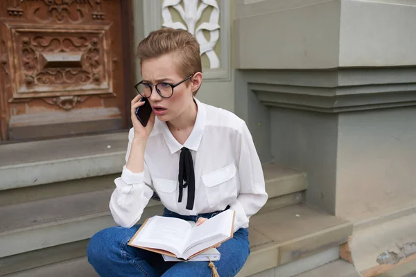 Studentin sitzt mit Buch in der Hand und Handy auf der Treppe neben dem Gebäude — Stockfoto