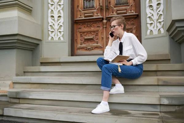 Studentin sitzt mit Buch in der Hand und Handy auf der Treppe neben dem Gebäude — Stockfoto