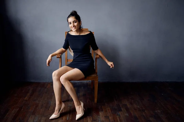 Όμορφη γυναίκα κάθεται σε μια καρέκλα σε ένα μαύρο φόρεμα δωμάτιο γκρι τοίχο μοντέλο χτένισμα — Φωτογραφία Αρχείου