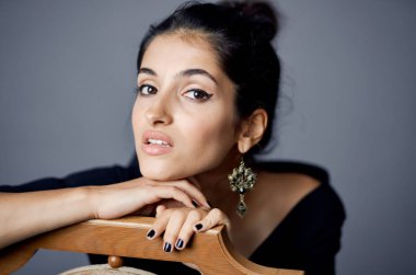 Siyah elbiseli seksi esmer gri bir odada tahta bir sandalyeye yaslanıyor.