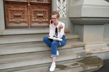 Kot gömlekli ve spor ayakkabılı bir kadın eğitim enstitüsü telefonunun yanındaki merdivenlerde oturuyor.