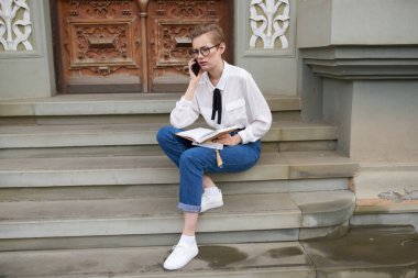 Kot gömlekli ve spor ayakkabılı bir kadın eğitim enstitüsü telefonunun yanındaki merdivenlerde oturuyor.