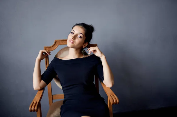 Mulher elegante em vestido preto senta-se na cadeira de madeira no quarto cinza — Fotografia de Stock