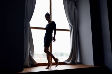 Arka plandaki perdelerin yanındaki pencere eşiğinde kısa elbiseli bir kadının silueti.