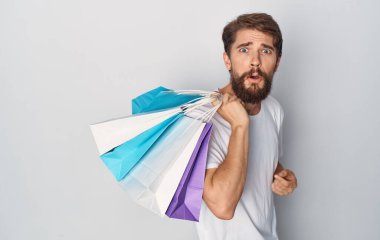 Beyaz tişörtlü sakallı adamın ellerinde paketlerle görüntüsü.