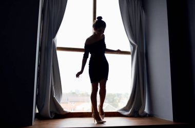 Arka plandaki perdelerin yanındaki pencere eşiğinde kısa elbiseli bir kadının silueti.