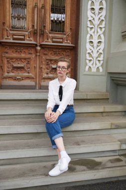 Spor ayakkabılı ve tişörtlü çekici bir kadın Eğitim Enstitüsü 'nün yanındaki basamaklarda oturuyor.