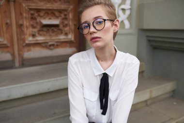 Kotlu ve gömlekli bir öğrenci binanın yanındaki basamaklarda oturuyor ve yüzünde gözlük var.