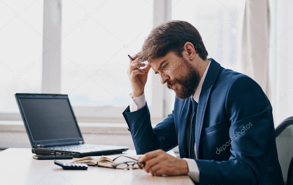 A man in a classic suit sits at a table and an open laptop office window a puzzled look