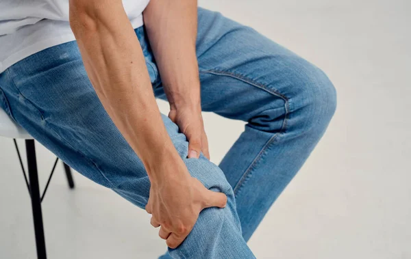 Чоловік в джинсах сидить на стільці і торкається ноги своїми руками обрізаний вид — стокове фото