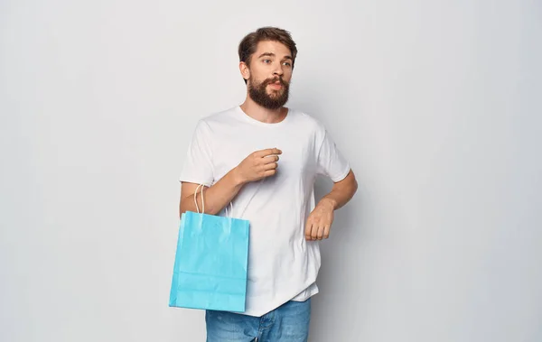 Homem barbudo em uma camiseta branca com um pacote em sua mão Compras divertido isolado fundo — Fotografia de Stock