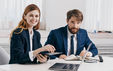 İş adamı ve kadın ofiste dizüstü bilgisayar ve not defteri modelleriyle masada oturuyorlar.