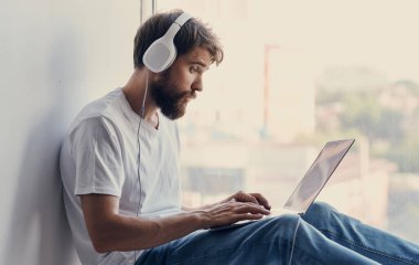 Bilgisayarı açık sakallı bir adam pencere eşiğinde ve kulaklıkları başında oturuyor.