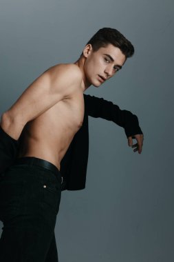 Koyu renk ceketli, çıplak, gövdesi gri arka planda bir adam.