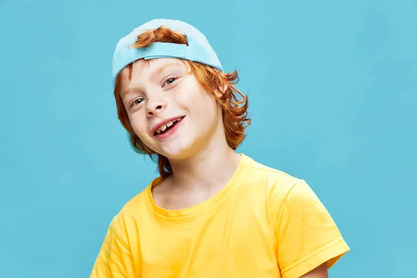 一个快乐的红头发男孩，穿着一件黄色的T恤，头上戴着一顶蓝色的帽子，带着童年时代的微笑 — 图库照片
