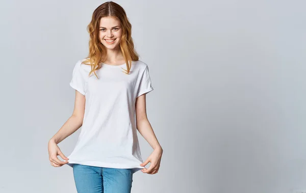 A beautiful woman in a white T-shirt and jeans smiles on a gray background and gestures with her hands — Stock Photo, Image