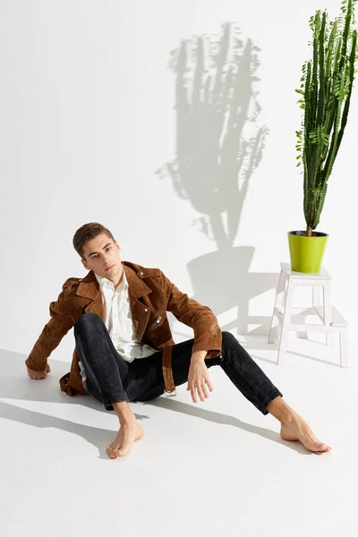 Un joven con una chaqueta marrón y pantalones negros se sienta en el suelo cerca de una flor en el interior — Foto de Stock