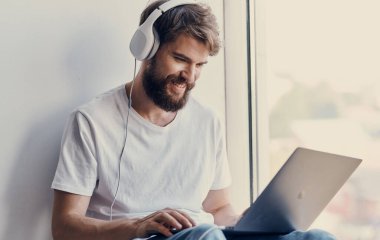 Bilgisayarı açık sakallı bir adam pencere eşiğinde ve kulaklıkları başında oturuyor.