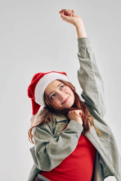 Una ragazza allegra in un cappello di Babbo Natale alzò la mano su uno sfondo leggero — Foto Stock