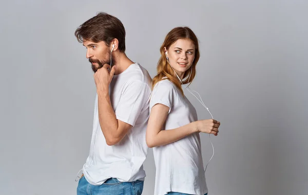 Hombre y mujer con auriculares cableados de pie con sus espaldas el uno al otro sobre un fondo gris en camisetas blancas — Foto de Stock