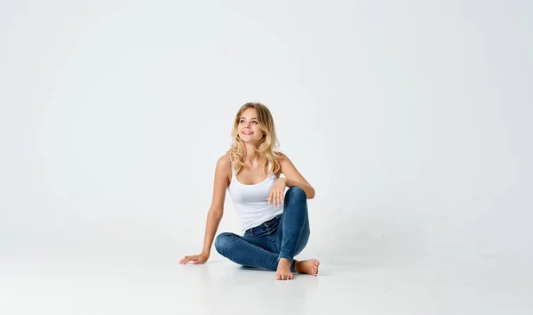 Piękna blondynka w dżinsach i biały t-shirt siedzi na podłodze wewnątrz i gesty z jej rąk — Zdjęcie stockowe