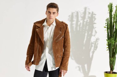 Beyaz gömlekli ve kahverengi ceketli bir adam. Arka planda, saksıda bir çiçeğin yanında.