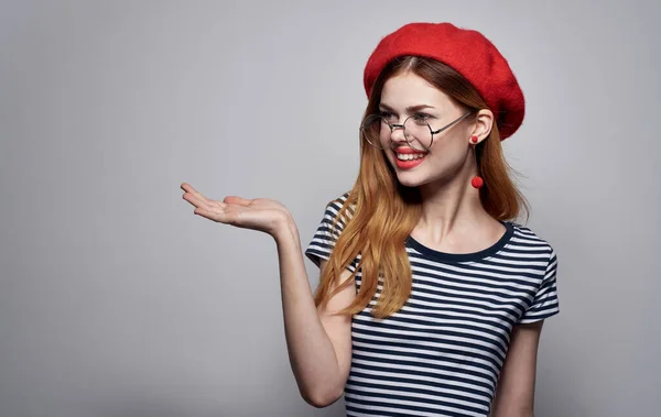 Femme émotionnelle dans des lunettes et dans un t-shirt rayé gestes avec ses mains chapeau rouge fond gris — Photo