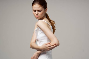 Beyaz tişörtlü, bej arka planda kızıl saçlı bir kadın. Elleri dirseğinde ağrı var.