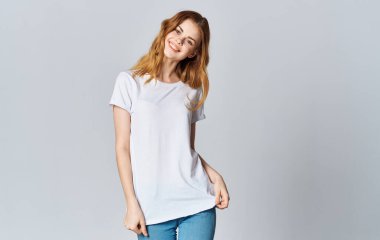 Beyaz tişörtlü ve kot pantolonlu güzel bir kadın gri arka planda gülümsüyor ve elleriyle hareket ediyor.