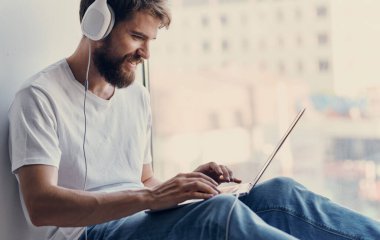 Kulaklıklı mutlu adam pencere kenarında oturuyor ve bilgisayar ekranına bakıyor.